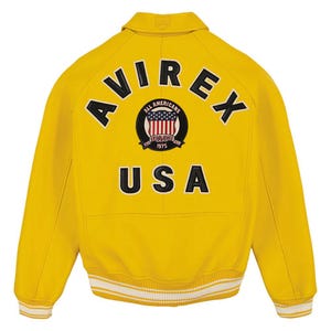 Pu&ograve; includere: Giacca in pelle gialla con la scritta "AVIREX" sulle spalle e "USA" sotto un emblema circolare. L'emblema presenta un disegno della bandiera americana e le parole "ALL AMERICANS ESTABLISHED 1975". La giacca ha un orlo a righe bianche e gialle.