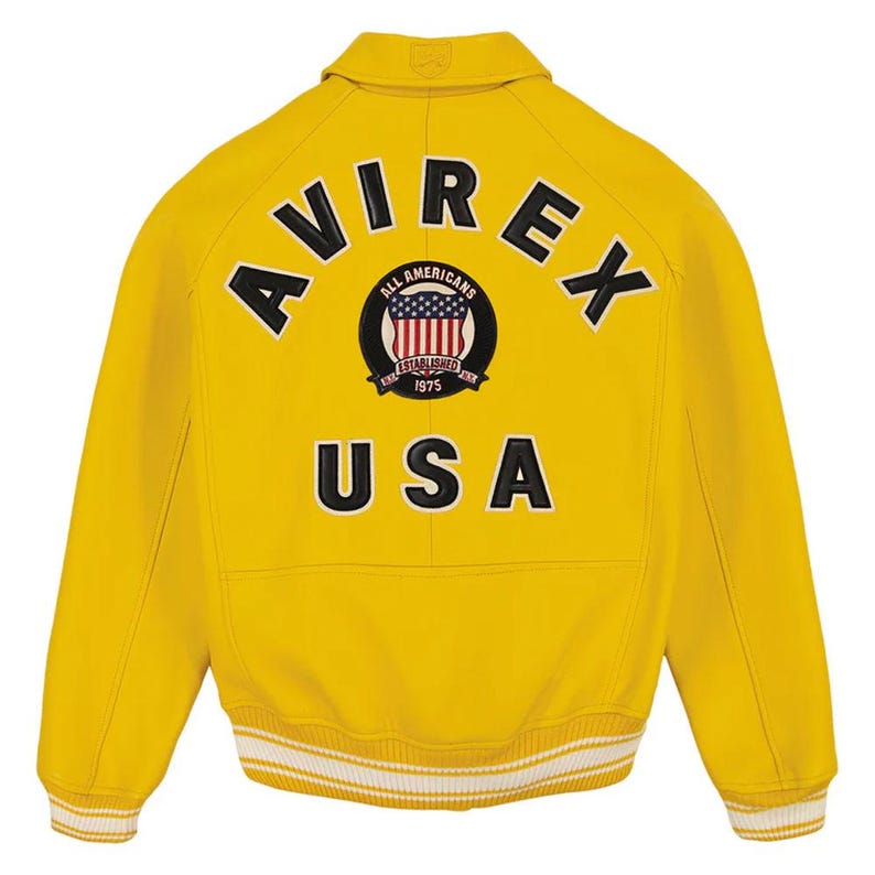 Pu&ograve; includere: Giacca in pelle gialla con la scritta "AVIREX" sulle spalle e "USA" sotto un emblema circolare. L'emblema presenta un disegno della bandiera americana e le parole "ALL AMERICANS ESTABLISHED 1975". La giacca ha un orlo a righe bianche e gialle.