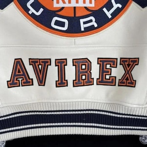 May include: Cream-colored varsity jacket with navy and orange accents. Features the text "KITH CORK" above "AVIREX" in orange lettering. The jacket has a striped navy and white ribbed hem.