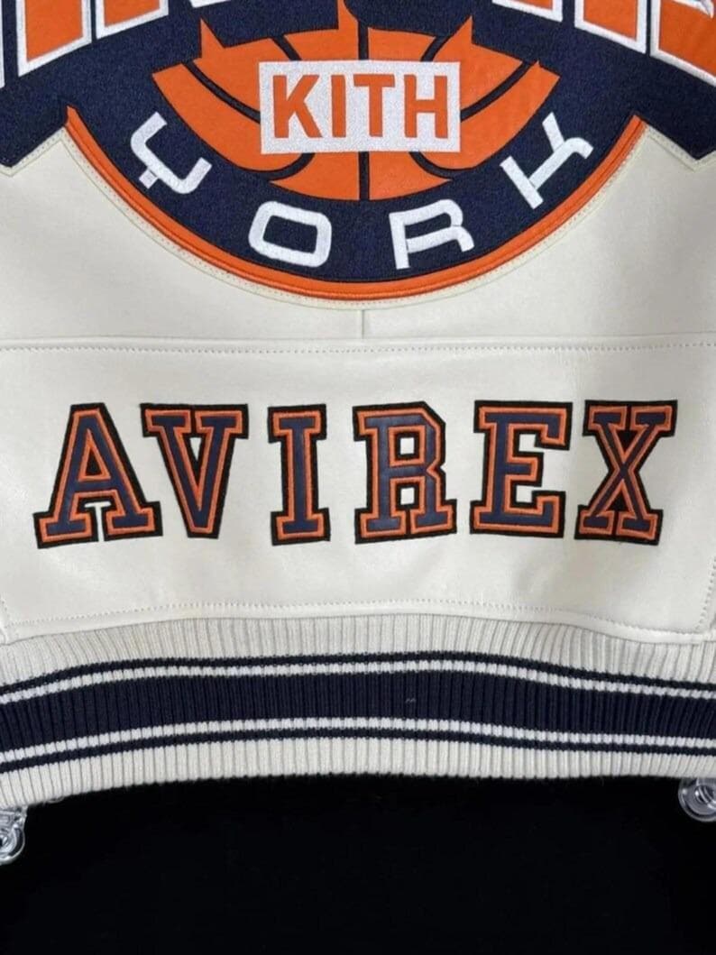 May include: Cream-colored varsity jacket with navy and orange accents. Features the text "KITH CORK" above "AVIREX" in orange lettering. The jacket has a striped navy and white ribbed hem.