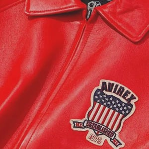 Pu&ograve; includere: Giacca in pelle rosso acceso con chiusura a zip e colletto classico. La giacca presenta una toppa sul petto con la scritta "AVIREX NY ESTABLISHED 1975" e un disegno della bandiera americana.