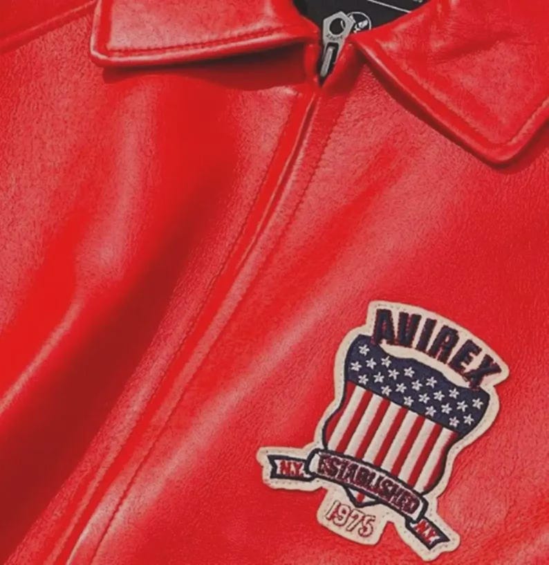 Pu&ograve; includere: Giacca in pelle rosso acceso con chiusura a zip e colletto classico. La giacca presenta una toppa sul petto con la scritta "AVIREX NY ESTABLISHED 1975" e un disegno della bandiera americana.
