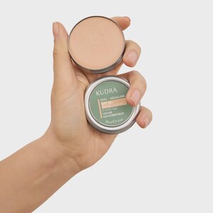 May include: A person holding two small, round, silver tins of sunscreen balm. One tin is open, revealing a light beige product. The other tin is closed, with a green label that reads "KUDRA ZINC + SQUALENE SPF 30 MINERAL SUNSCREEN FILTER".