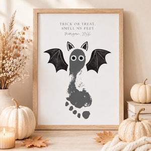 Op de afbeelding: Een ingelijste print met een grijze voetafdruk die is veranderd in een vleermuis met zwarte vleugels en twee ogen. De tekst luidt: TRICK OR TREAT, SMELL MY FEET, Morgan, 20XX. Het kunstwerk zit in een lijst van lichtgekleurd hout.