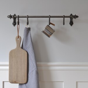 May include: A metal kitchen rack with hooks, holding a wooden chopping board, a grey kitchen towel, and a silver jug.