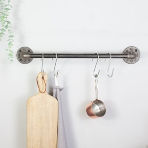 May include: A black metal pipe rack with five hooks hanging from it. A wooden chopping board, a grey kitchen towel, and a copper measuring cup are hanging from the hooks.