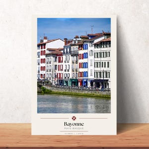 Peut inclure: Une impression verticale montrant une rangée de bâtiments à plusieurs étages aux façades blanches avec des volets rouges, bleus ou verts le long d'une rive. Sous l'image, le texte indique Bayonne PAYS BASQUE avec les coordonnées 43.488 N | -1.475 W.