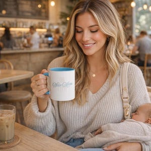 May include: A white coffee mug with a blue interior and handle, featuring the text "The New Mom Club" in light blue script. The mug is held by a person in a light-colored sweater.