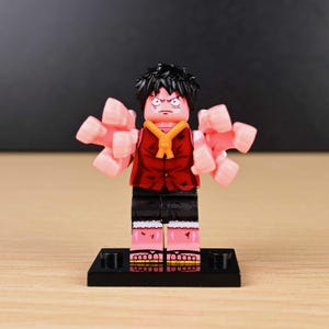 May include: A Lego-style figure of a character with black hair, a red vest, and black shorts. The figure has pink fists extending from its arms and is standing on a black base. The character has an angry expression.