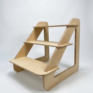 Peut inclure: Étagère en bois à trois niveaux, avec une finition de couleur claire. L'étagère a une conception en gradins, chaque niveau étant soutenu par des supports latéraux inclinés. Idéal pour exposer des objets.