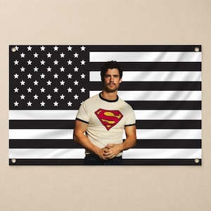 May include: A rectangular wall banner featuring a black and white American flag design with metal grommets in each corner. Centered on the flag is a photograph of a person with dark hair wearing a cream-colored ringer t-shirt with a red and yellow Superman logo on the chest.