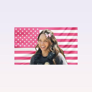 May include: A smiling person with long brown hair and a pink bow in their hair. They are wearing a dark gray shirt and a black vest. Behind them is a pink American flag.