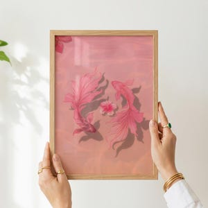 Op de afbeelding: Een ingelijste kunstdruk met twee roze koi-vissen die zwemmen in een roze waterlandschap. Een roze bloem zweeft tussen de vissen. Het kunstwerk wordt vastgehouden door handen met gouden ringen.