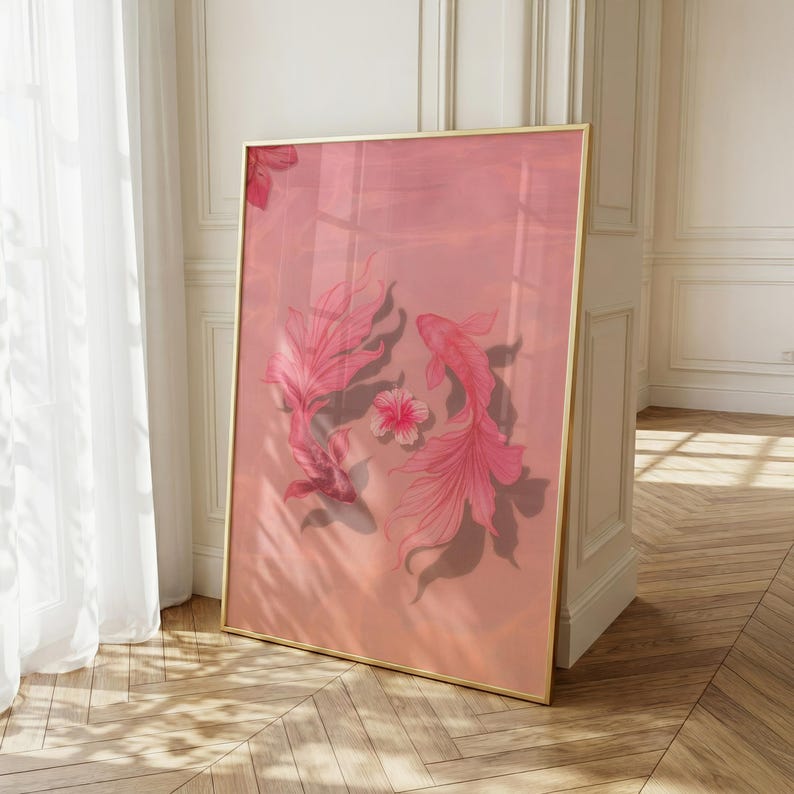 Op de afbeelding: Een ingelijste kunstdruk met twee roze koi-vissen en een bloem op een roze achtergrond. Het kunstwerk heeft een gouden lijst en leunt tegen een witte muur. De afbeelding wordt verlicht door natuurlijk licht.