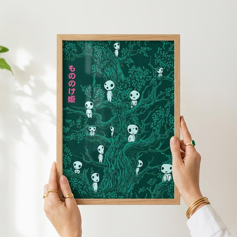 May include: A framed art print featuring a dark green illustration of a tree with numerous white, spirit-like figures. The Japanese text "もののけ姫" is printed in pink on the left side. The print is held by hands with gold rings and bracelets.
