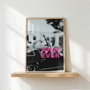 May include: A framed black and white photograph with a person raising their arms in a car, and the pink text "WHATEVER". The image is in a light wood frame and rests on a wooden shelf against a white wall.