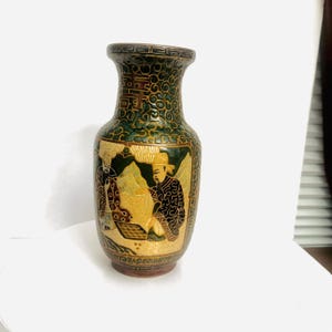 May include: A decorative ceramic vase featuring a dark green background with intricate gold-toned patterns and a central panel depicting two figures engaged in a board game. The neck of the vase displays a repeating geometric border design.