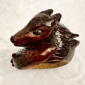 May include: A dark brown glazed ceramic ring featuring a sculpted animal head with pointed ears and a smooth, glossy finish. The band of the ring has a speckled, light tan, unglazed texture on the interior, contrasting with the dark exterior.