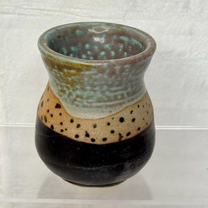 May include: A small ceramic cup with a rounded body and a flared rim. The exterior features a dark brown base, a middle section of speckled tan, and a top section glazed in a mottled light blue and grey finish with a smooth, glossy texture.