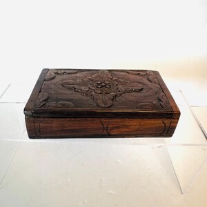 May include: A rectangular dark brown wooden box featuring a hand-carved floral motif on the lid. The lid displays a central flower design surrounded by intricate leaf patterns and dotted textures, while the sides have simple carved lines.