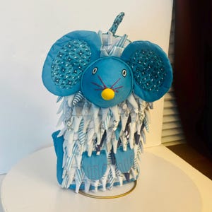 May include: A plush mouse toy featuring a bright blue face with a yellow nose, embroidered eyes, and pink whiskers. The body is covered in layered, shaggy fabric strips in shades of blue, white, and grey, with large, patterned blue ears.