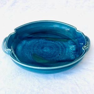 May include: A handmade, oval-shaped ceramic baking dish with a glossy, deep teal glaze. The dish features a scalloped rim and integrated handles. The interior shows variations in color, with hints of green and lighter blue, creating a marbled effect. The dish is designed for oven use.