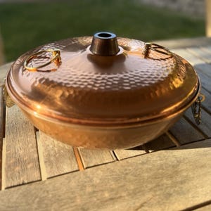 May include: A round, hammered copper serving dish with a matching lid. The lid features a central dark metal knob and two gold-toned ring handles on opposite sides. The exterior surface has a textured, dimpled finish throughout the metal body.
