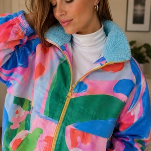 May include: A zip-up jacket featuring a vibrant, abstract pattern in shades of pink, blue, and green. The jacket has a light blue sherpa collar, long sleeves with elastic cuffs, and a gold-toned metal zipper closure down the center front.