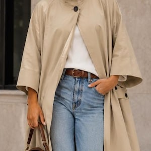 May include: A beige trench coat with a high collar and a single dark button closure, worn over a white ribbed top and light blue denim jeans with a brown leather belt. A small brown leather handbag is held at the side of the long coat.