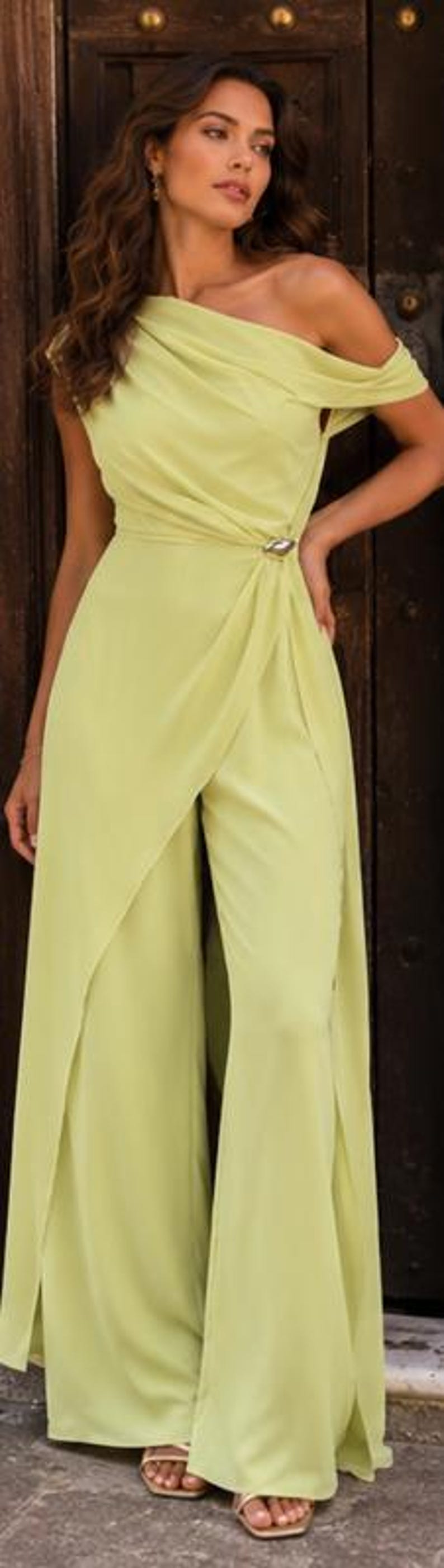 May include: A light lime green jumpsuit featuring an asymmetrical off-the-shoulder neckline with draped fabric across the bodice. The waist is accented with a silver-toned metallic detail, and the wide-leg trousers include a front overlay panel.