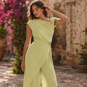 May include: A light lime green jumpsuit featuring a draped cowl neckline, short cap sleeves, and a fitted waist accented with a metallic circular embellishment. The garment has a wrap-style front overlay that cascades over wide-leg trousers.