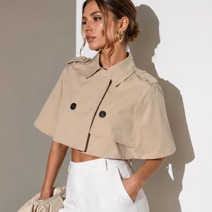 May include: A beige cropped trench cape featuring a classic collar, shoulder epaulets, and two prominent black buttons on the front. The garment has wide, short sleeves and a structured, flared silhouette that ends above the waistline.