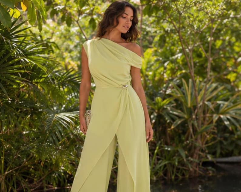 May include: A light lime green one-shoulder jumpsuit featuring a draped bodice, a fitted waist with a decorative silver-toned brooch detail, and wide-leg trousers with an overlapping front panel design that creates a fluid, layered silhouette.