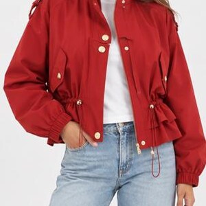May include: A vibrant red cropped jacket featuring a high collar, gold-toned buttons, and a drawstring waist with adjustable toggles. The jacket has long sleeves with elastic cuffs, shoulder epaulets, and a front zipper closure over a white shirt.