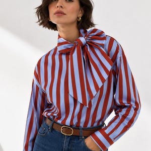 Op de afbeelding: Een gestreept overhemd in rood en lichtblauw met een grote strikdetail bij de hals. Het overhemd wordt gedragen met een bruine riem en een blauwe jeans. De riem heeft een gouden gesp. Afmetingen in centimeters.