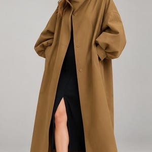 May include: A long, tan-colored trench coat featuring a high, structured collar, a button-front closure, and voluminous sleeves with buttoned cuffs. The coat is worn over a simple black dress with a front slit and black high-heeled pumps.
