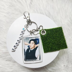 May include: A keychain featuring a rectangular acrylic charm with a photograph of a person in a white shirt and black vest, a string of white letter beads spelling ONLYSANS with a blue heart, and a square patch of green artificial grass.