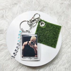 May include: A keychain featuring a rectangular acrylic charm with a photograph of a person in a black tank top taking a mirror selfie, a square piece of green artificial grass, and a string of white beads spelling out the text ONLY CHANS.