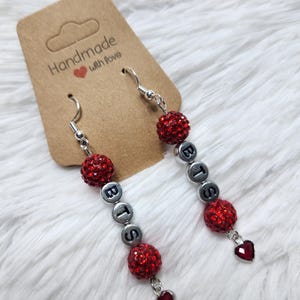 May include: Dangle earrings with silver hooks, featuring red rhinestone-covered beads and silver letter beads spelling "BTS". Each earring has a small red heart charm. The earrings are displayed on a white, fluffy surface.