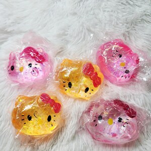 May include: Five individual squishy toys shaped like Hello Kitty heads, each encased in clear plastic packaging. Two toys are yellow with red bows, and three are pink with red bows, all featuring glittery details and black facial features.
