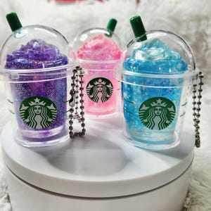 May include: Three miniature clear plastic cups with dome lids and green straws, each containing glittery purple, pink, or blue liquid. Each cup features a green circular Starbucks logo on the front and includes a silver metal ball chain keychain.