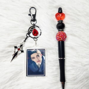Può includere: Una penna nera con una parte superiore decorativa con perline rosse e nere e un accento a forma di cuore. È presente anche un portachiavi con una foto, una gemma rossa e un ciondolo a forma di croce.