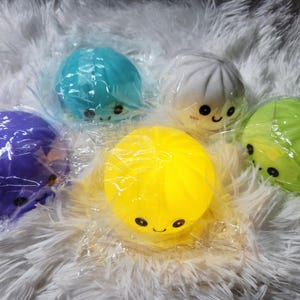 May include: Five round, squishy dumpling-shaped toys in purple, blue, white, yellow, and green. Each toy features a simple face with two black eyes and a small smile, and is individually wrapped in clear plastic packaging for protection.