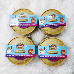 May include: Four round, tan, woven-textured plastic containers, each wrapped in a blue and pink paper band labeled Squishy Bun One Set Sugar Edition. The packaging features a cartoon bun character, a mystery basket graphic, and text for ages 3+.