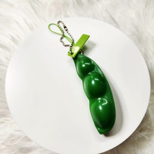 May include: A green plastic pea pod keychain featuring three rounded, protruding pea shapes within the pod. The item includes a small, light green plastic tab at the top attached to a silver-colored metal ball chain and a thin green cord loop.