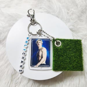 May include: A keychain featuring a rectangular acrylic charm with a photograph of a shirtless person, a strand of white beads spelling ONLYCHANS with a blue heart, and a square piece of green artificial grass attached by a silver metal ring.