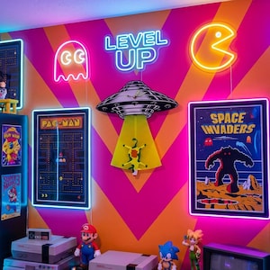 May include: A vibrant gaming room wall featuring neon signs of a ghost, the text LEVEL UP, a Pac-Man character, and the word ARCADE. The wall displays framed posters of Pac-Man and Space Invaders, alongside a UFO wall clock and shelves with figures.