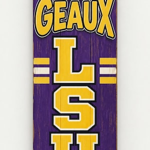 May include: A vertical wooden sign with a purple background featuring a tiger head at the top, the yellow text GEAUX LSU stacked vertically in the centre, and a brown football resting in green grass at the bottom with white accent stripes.