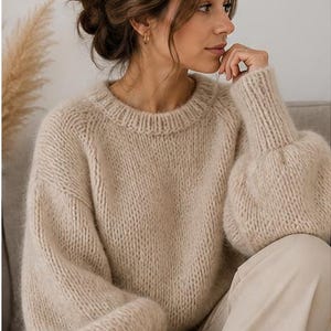 Puede incluir: Un suéter de punto beige de gran tamaño con cuello redondo y mangas largas y voluminosas. El suéter se combina con pantalones de color claro. La imagen muestra a una mujer que lleva el suéter, sentada en el interior.