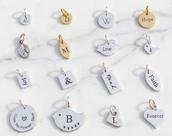 May include: A collection of sixteen silver and gold-toned metal charms in various shapes including circles, hearts, birds, and rectangles. Each charm is engraved with letters, names, or words such as Hope, Love, Joy, Faith, and Forever.